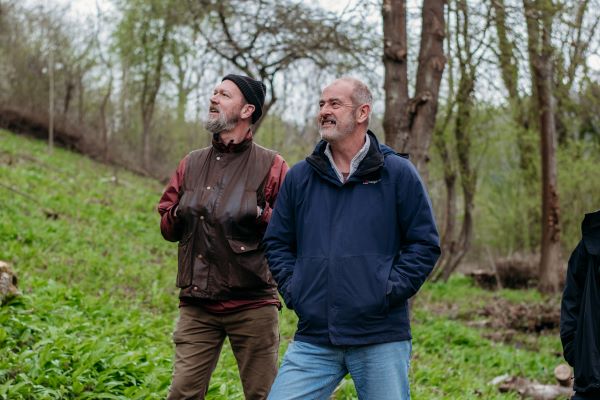 Two people standing at the edge of a woodland. They are looking up at the trees and smiling.